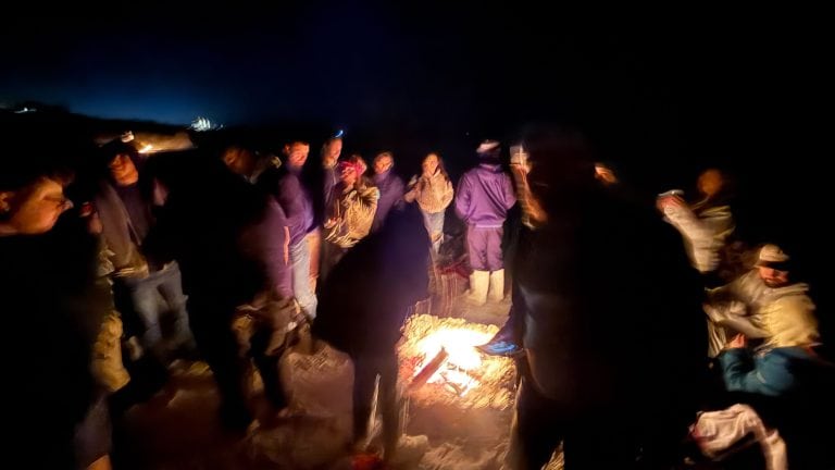 People are gathered around a beach bonfire at night.
