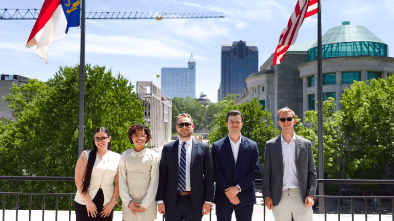 A group of five people in business attire stand outdoors with city buildings in the background, flanked by two flags.